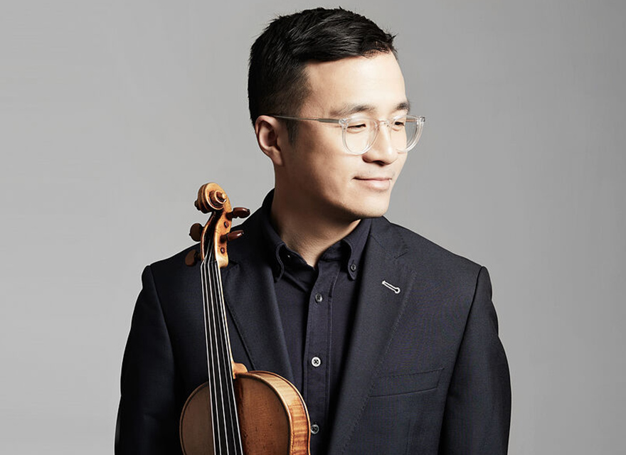 The image shows violinist Andrew Wan with short black hair and glasses, wearing a dark suit and holding a violin. He is looking slightly to his right with a calm and confident expression. The background is a plain, light grey color.