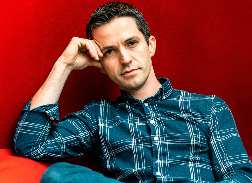 The image shows composer Ian Cusson with short dark hair wearing a dark green plaid shirt. He is sitting against a red background and resting his head on his right hand, with his elbow propped up. He has a serious expression on his face.