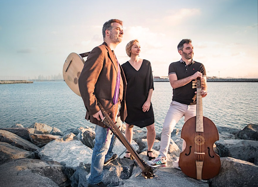 This image shows three members of the French Baroque music ensemble Faenza standing on rocky terrain with a cityscape in the background, including the Burj Khalifa. The date "Oct 19" is prominently displayed in the lower left corner. The person on the left holds a lute, the person in the center stands with their hands at their sides, and the person on the right holds a large stringed instrument called a viola da gamba.