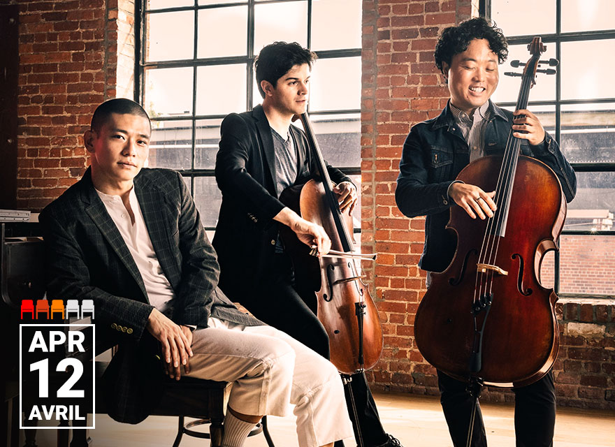 This image shows the NYC-based trio Empire Wild. The three members are posing with their instruments in a stylish, rustic indoor setting with brick walls and large windows. Two of them are holding cellos, while the third is seated, leaning back in a relaxed manner. The atmosphere is casual yet professional. The date "Apr 12" is prominently displayed in the lower left corner.