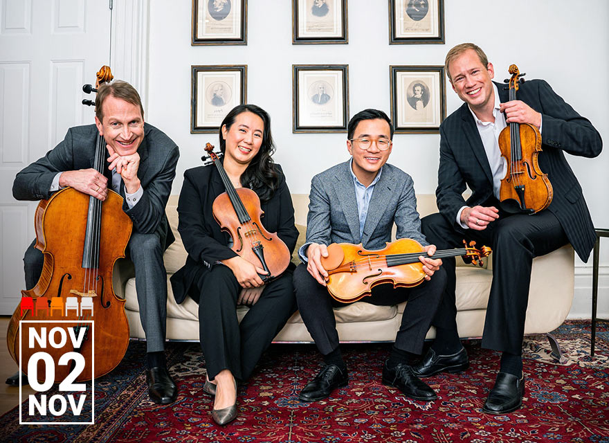 This image shows the members of the New Orford String Quartet sitting on a couch and smiling. They are dressed in formal attire. The quartet members are holding their respective string instruments: two violins, a viola, and a cello. The background features framed pictures on the wall. The date "Nov 02" is prominently displayed in the lower left corner.