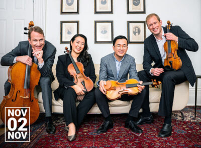 This image shows the members of the New Orford String Quartet sitting on a couch and smiling. They are dressed in formal attire. The quartet members are holding their respective string instruments: two violins, a viola, and a cello. The background features framed pictures on the wall. The date "Nov 02" is prominently displayed in the lower left corner.\