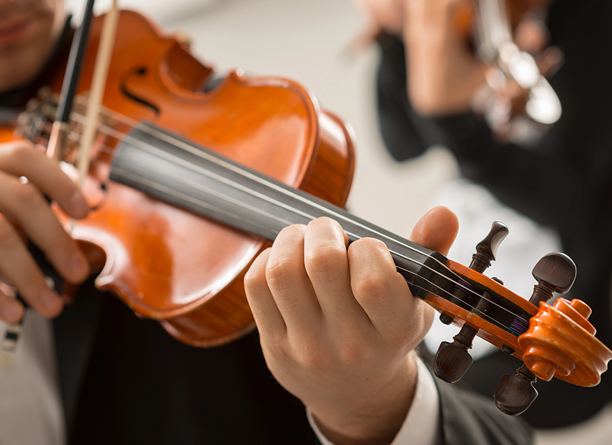 A close-up of a violinist's hand skillfully pressing the strings of a brightly polished violin, with another violinist visible in the blurred background, both dressed formally.